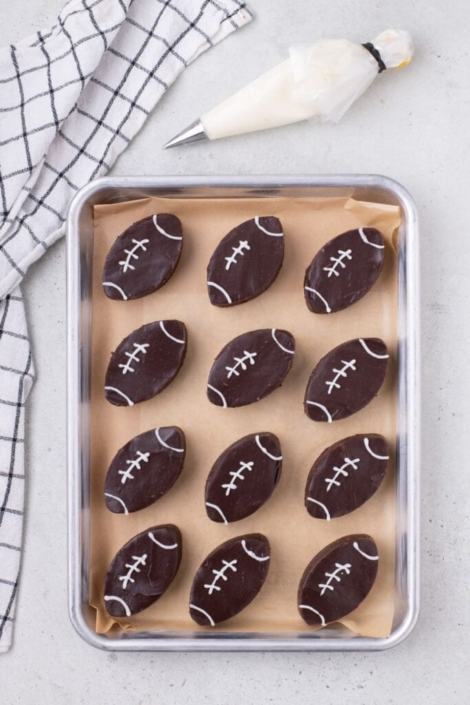 A parchment lined baking sheet with football brownies on it. A bag of white icing is above the sheet.