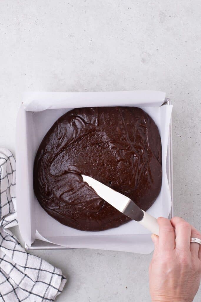 Brownie batter is smoothed into a baking pan with an offset spatula.