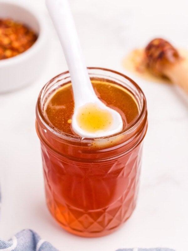 Completed hot honey sauce in a small mason jar with a white ceramic spoon resting on the surface of the sauce.