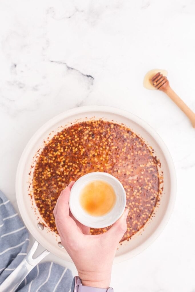 Pan with red chili flakes and honey. A hand is holding a small pinch bowl with apple cider vinegar above the pan.