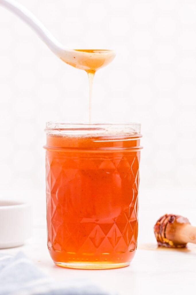 Hot honey in a small mason jar. A white ceramic spoon is lifting a scoop of honey out of the jar.