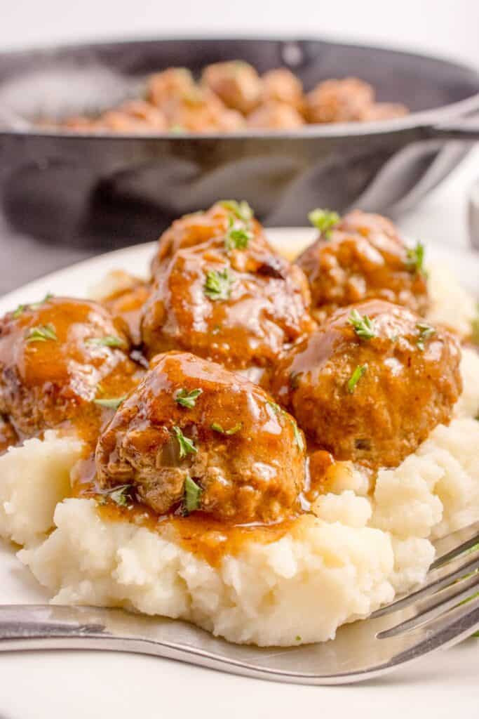 Meatballs with gravy on a bed of mashed potatoes on a white dinner plate with a fork.