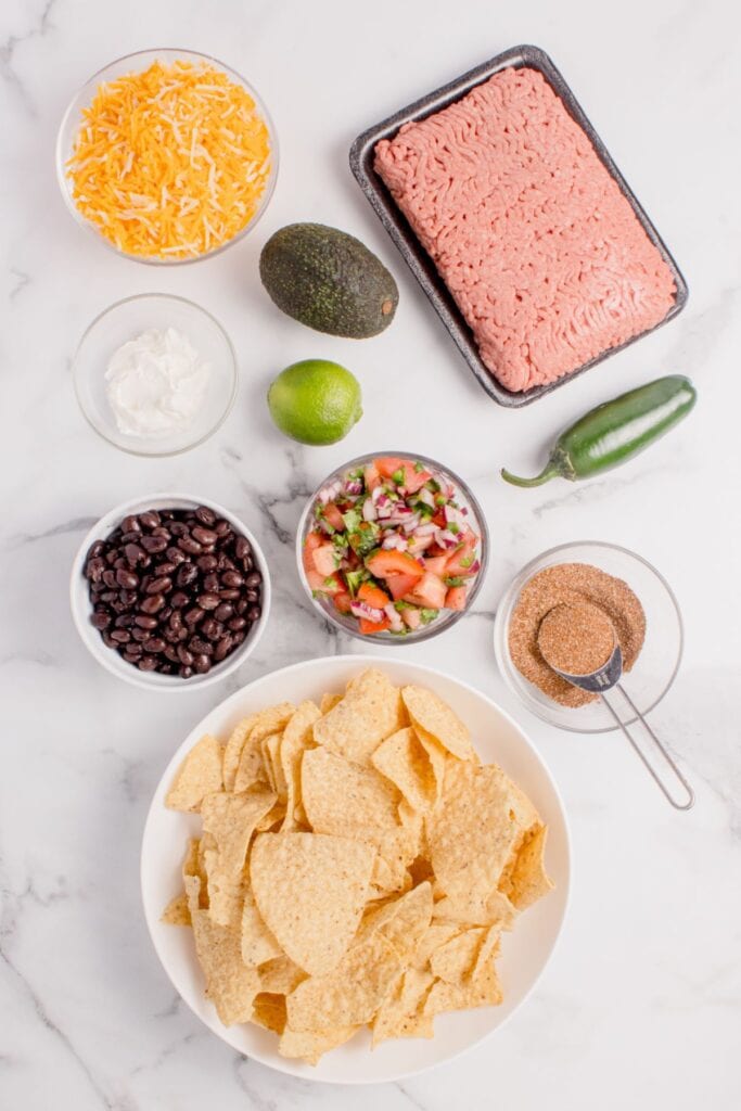 Ingredients for oven nachos: ground beef, shredded cheese, avocado, sour cream, lime, jalapeno, black beans, pico de gallo, taco seasoning, and tortilla chips.
