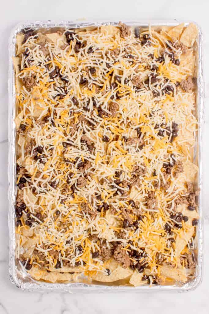 Sheet pan loaded with tortilla chips, ground beef, black beans, and shredded cheese.