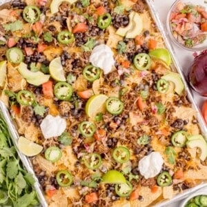 Completed oven nachos on an aluminum foil covered baking sheet surrounded by nacho toppings in bowls.