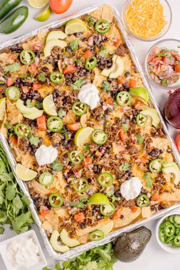 Completed oven nachos on an aluminum foil covered baking sheet surrounded by nacho toppings in bowls.