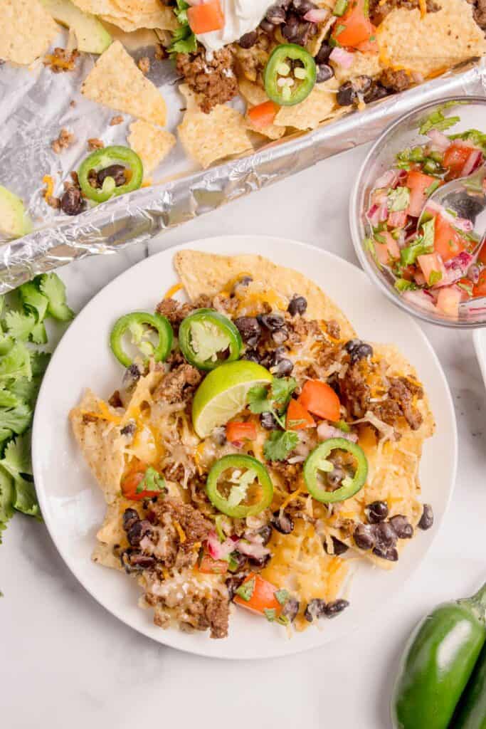 Completed oven nachos on a white dinner plate next to an aluminum foil covered baking sheet with the nachos recipe.