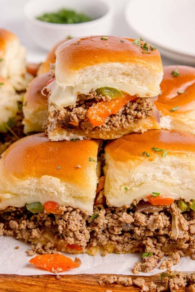 Philly cheesesteak sliders stacked on a parchment covered wooden cutting board.