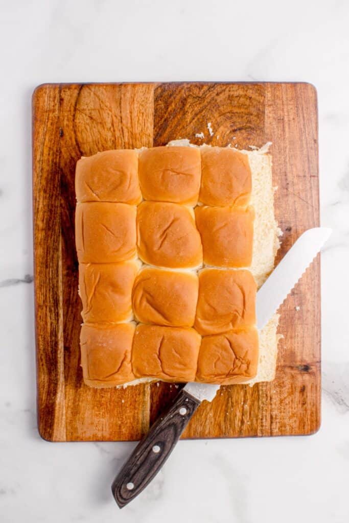 Hawaiian rolls being sliced in half on a wooden cutting board.