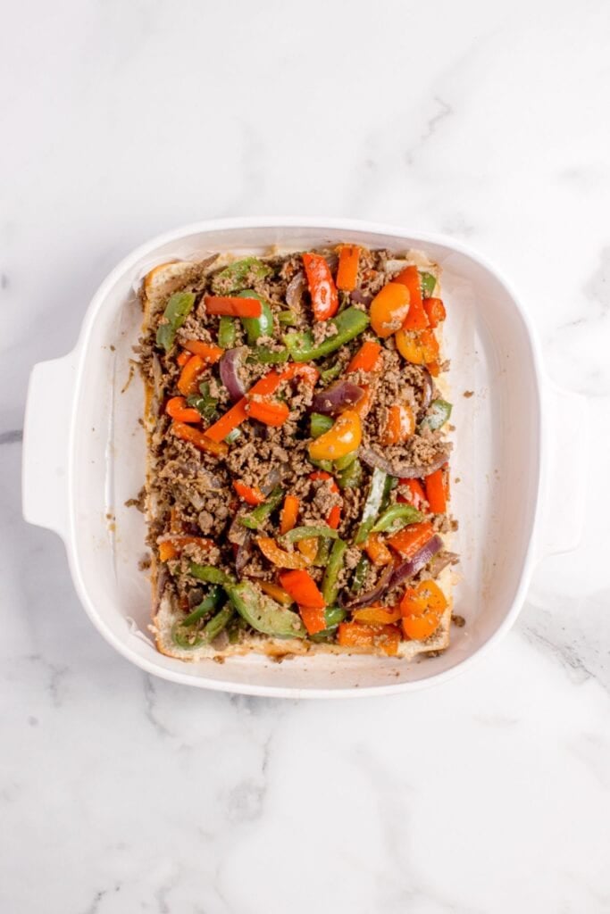 A white baking pan with the bottom half of hawaiian rolls and a layer of beef, peppers, and onions on it.