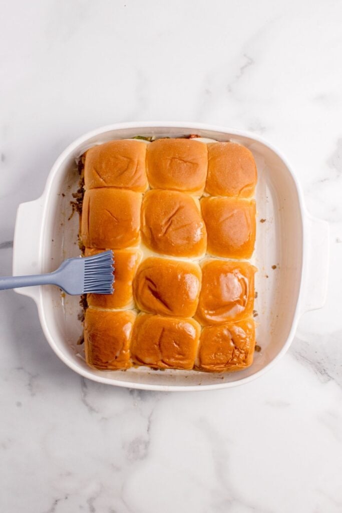White baking pan with unbaked cheesesteak sliders being brushed with butter.