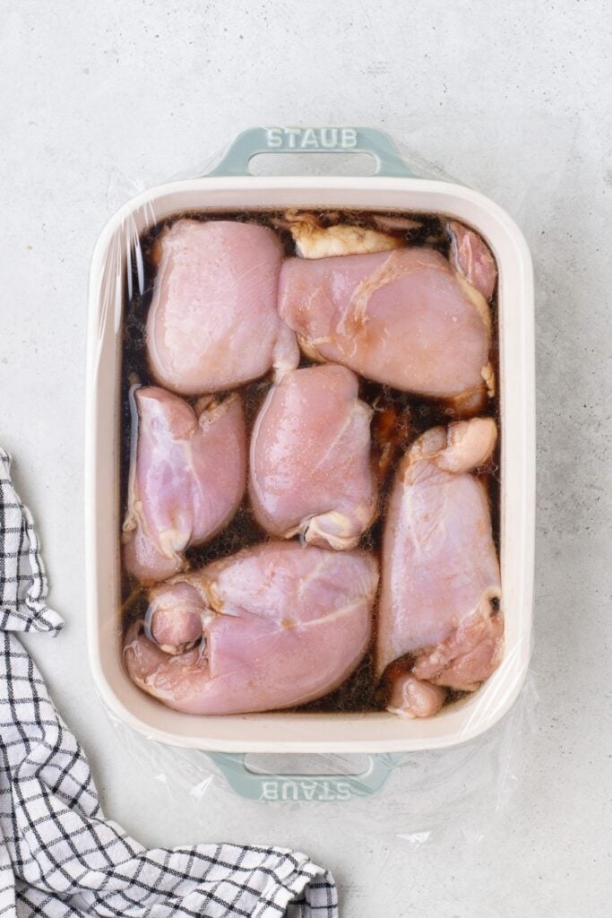 Marinating chicken thighs in a baking dish with plastic wrap over the top.