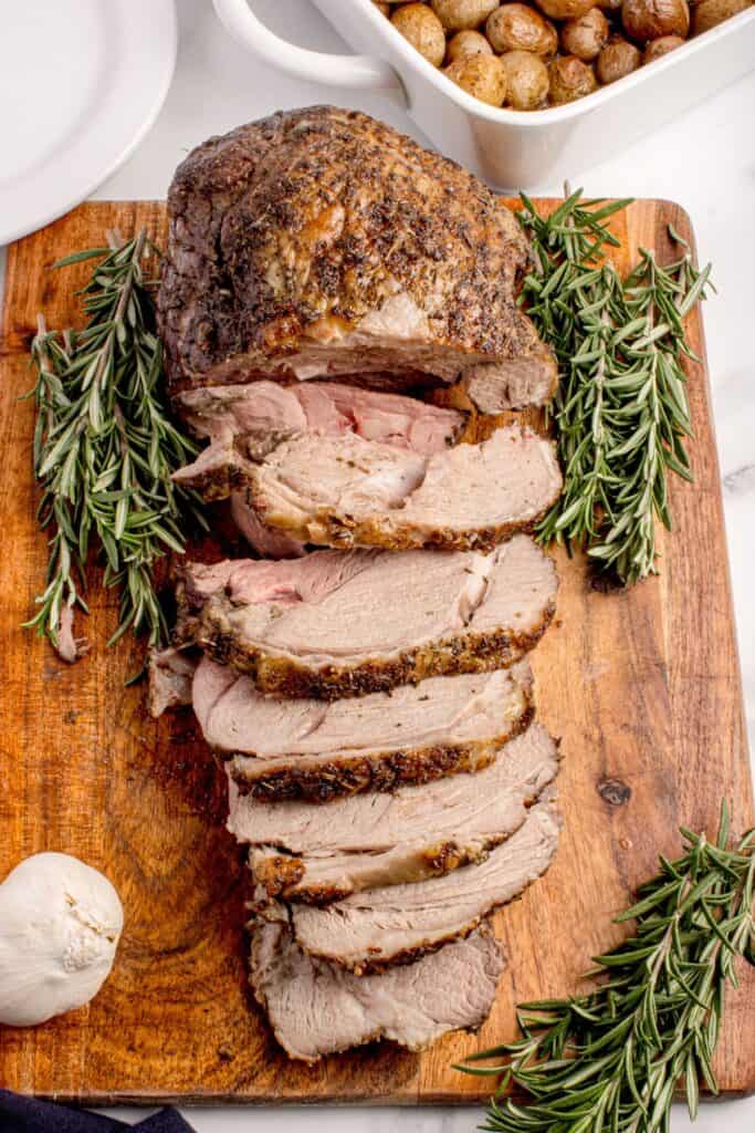 Sliced leg of lamb on a wooden cutting board, garnished with rosemary.