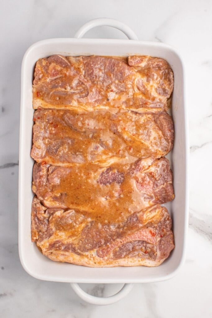 A white baking dish with marinated pork steaks lined in it.