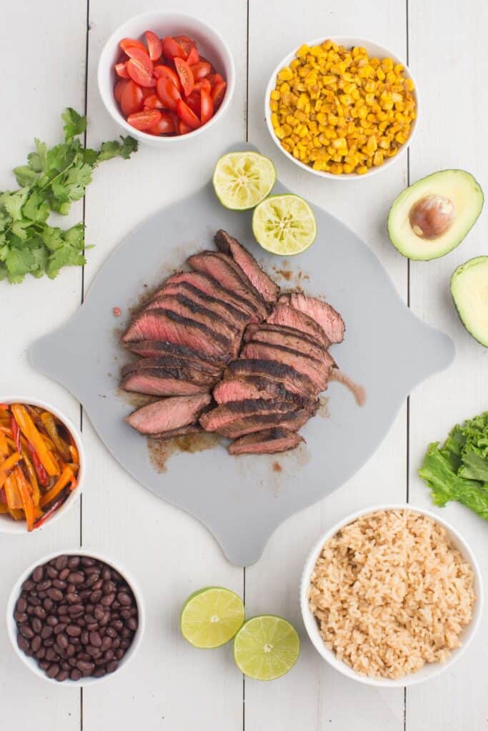 Sliced steak on a gray cutting board. Steak is surrounded by all the ingredients for steak bowls.