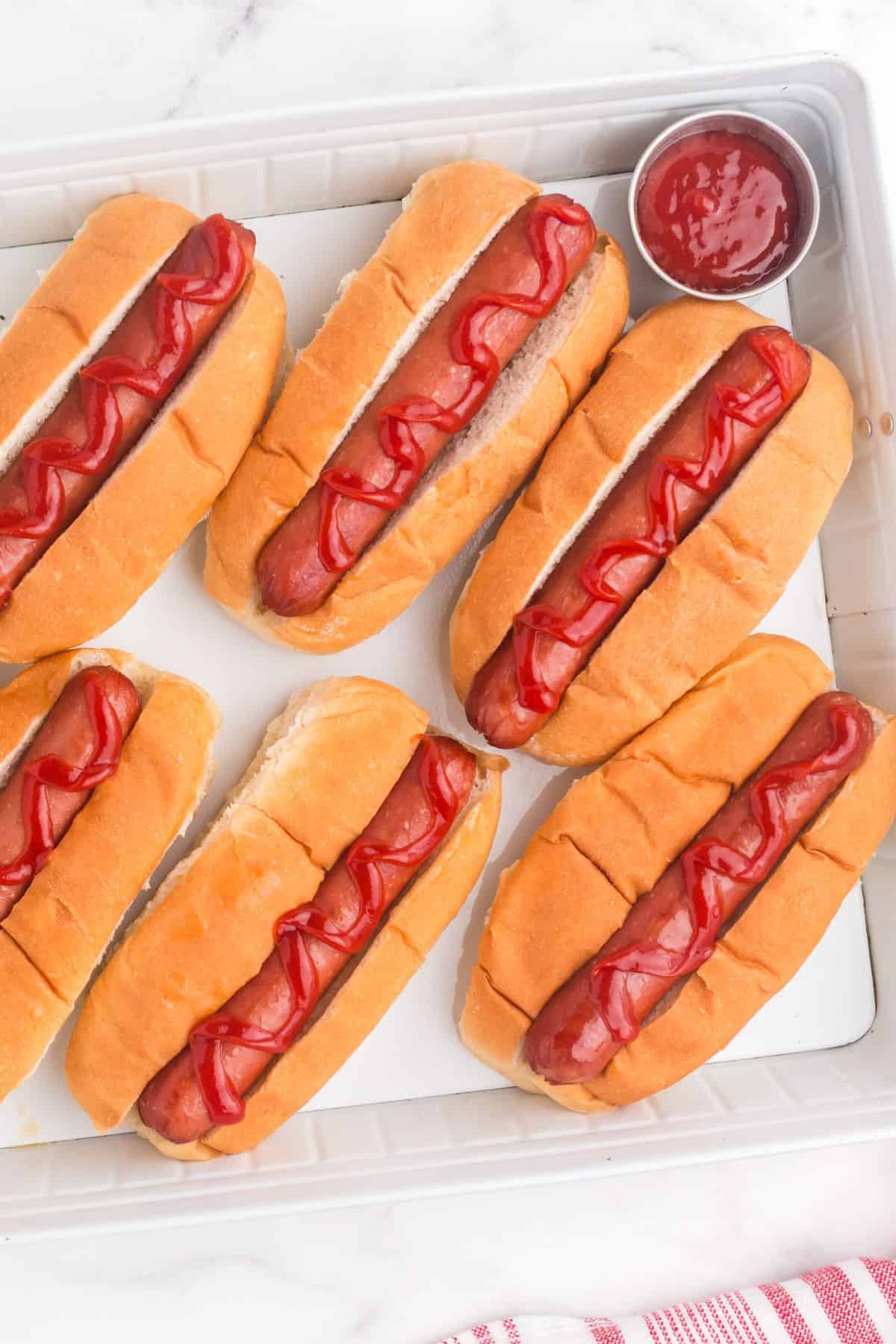 Several completed hot dogs on a tray with a little cup of ketchup.