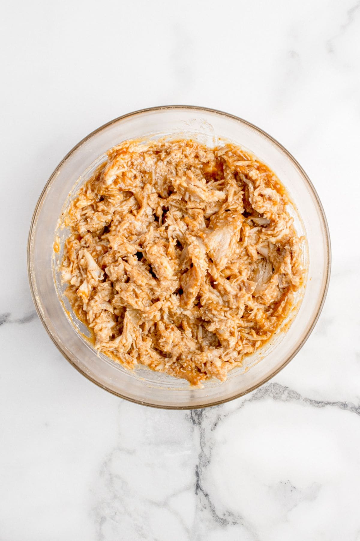 Glass mixing bowl with shredded chicken and BBQ sauce in it.