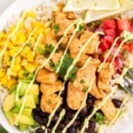Finished burrito bowl in a white bowl with rice, seasoned chicken, black beans, corn, avocado, pico de gallo, lime wedges and avocado sauce.