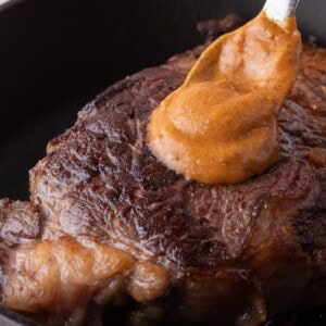 Cooked steak in a cast iron pan. A spoon is dolloping a serving of cowboy butter on top.