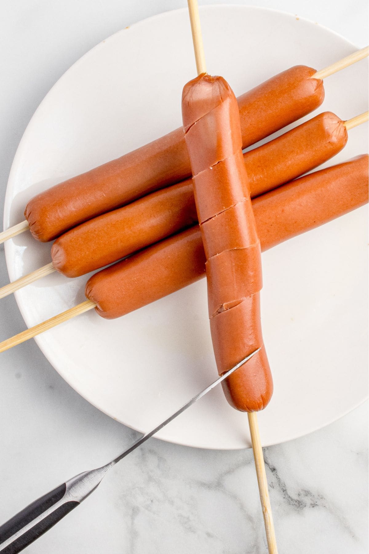 A knife is slicing the hot dog on the skewer.