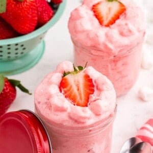 Two servings of strawberry fluff in small glass jars. A colander of strawberries sits behind the jars.