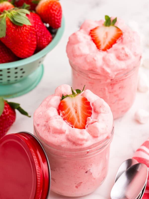 Two servings of strawberry fluff in small glass jars. A colander of strawberries sits behind the jars.
