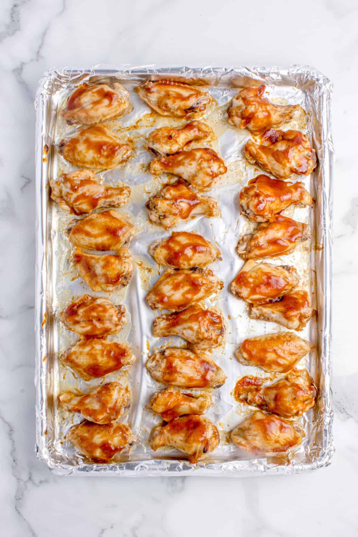 Par baked chicken wings on a foil lined baking sheet. Wings have a coat of BBQ sauce on them.