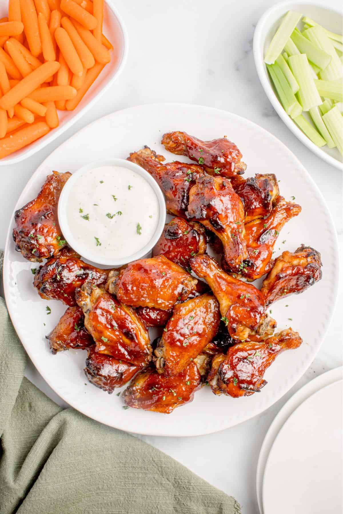 Chicken wings on a white serving platter with a small bowl of creamy dip. Platter is flanked by bowls of baby carrots and celery pieces.