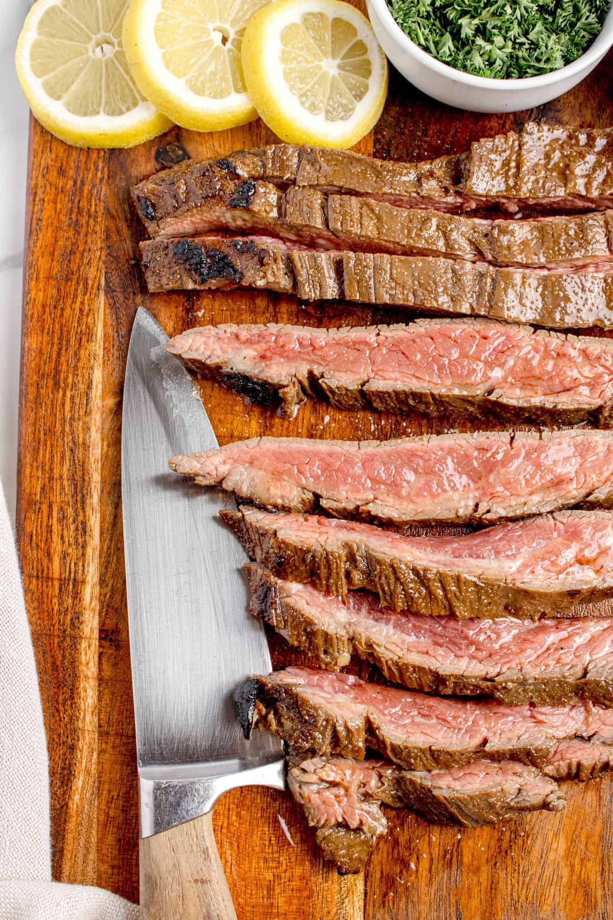 Cooked and sliced flank steak on a wooden cutting board. A knife is placed next to the sliced steak.