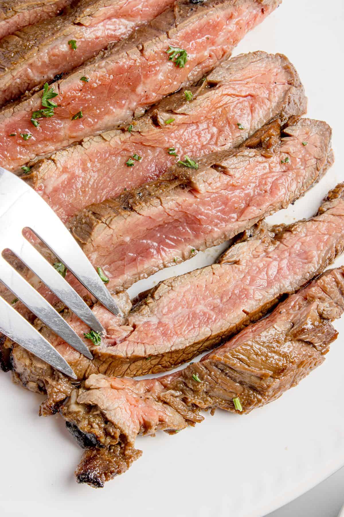 Cooked flank steak sliced and on a white dinner plate. A fork is piercing a slice of steak.