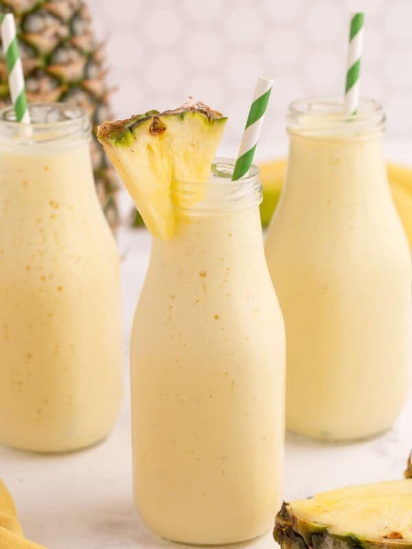 Three milk style bottles filled with pineapple smoothie. Each glass has a white and green striped straw in it and one glass has a small pineapple wedge on the rim.