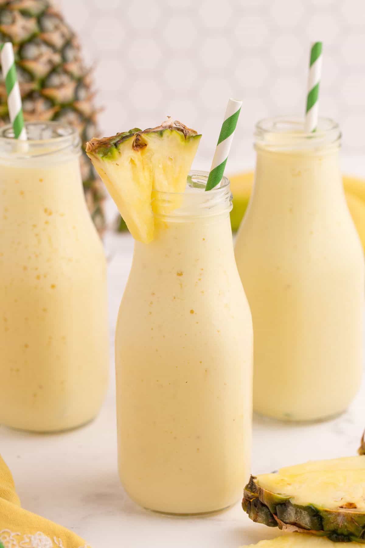 Three milk style bottles filled with pineapple smoothie. Each glass has a white and green striped straw in it and one glass has a small pineapple wedge on the rim.