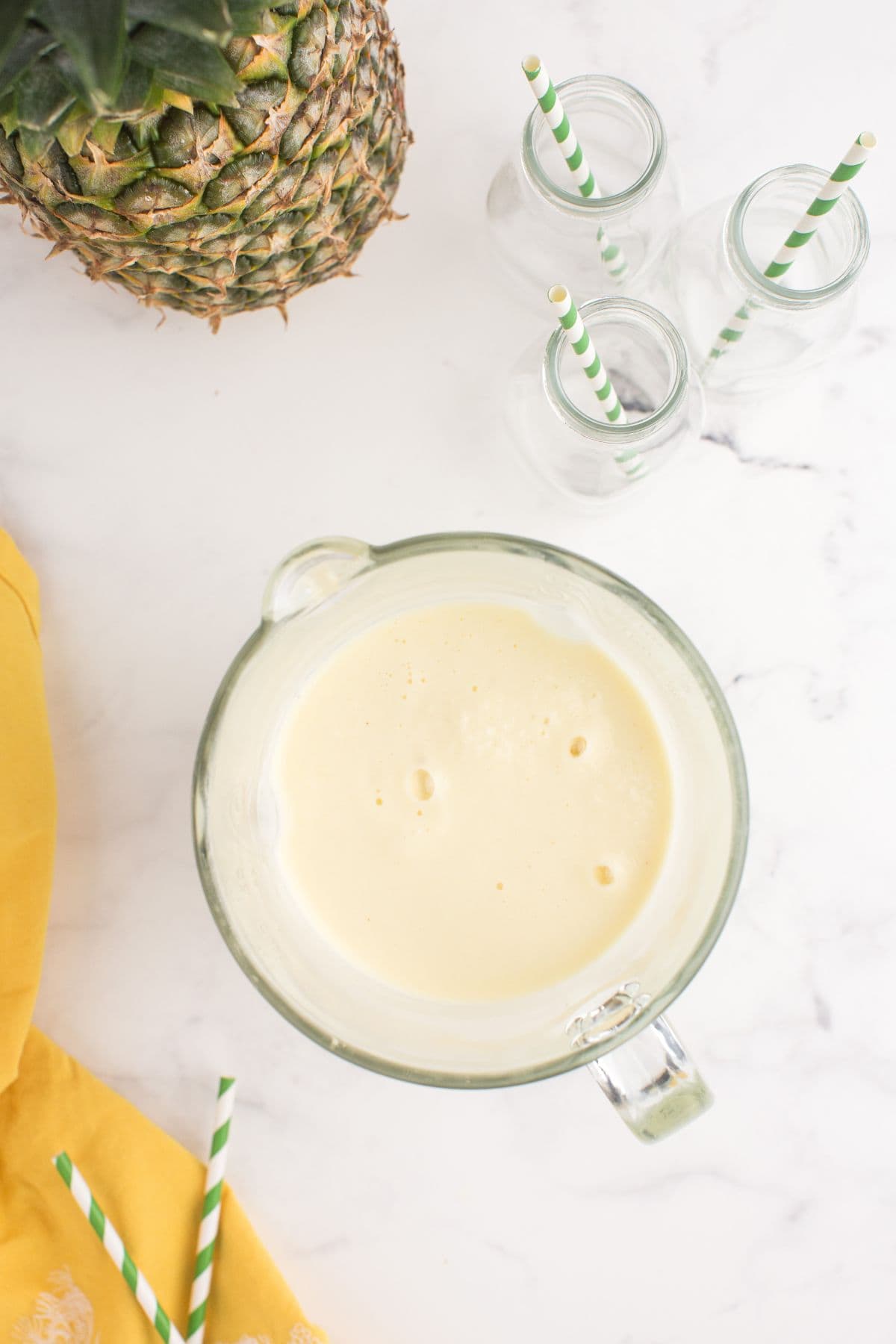 Glass blender with blended pineapple smoothie in it. Three milk style glasses with green and white straws are next to the blender.