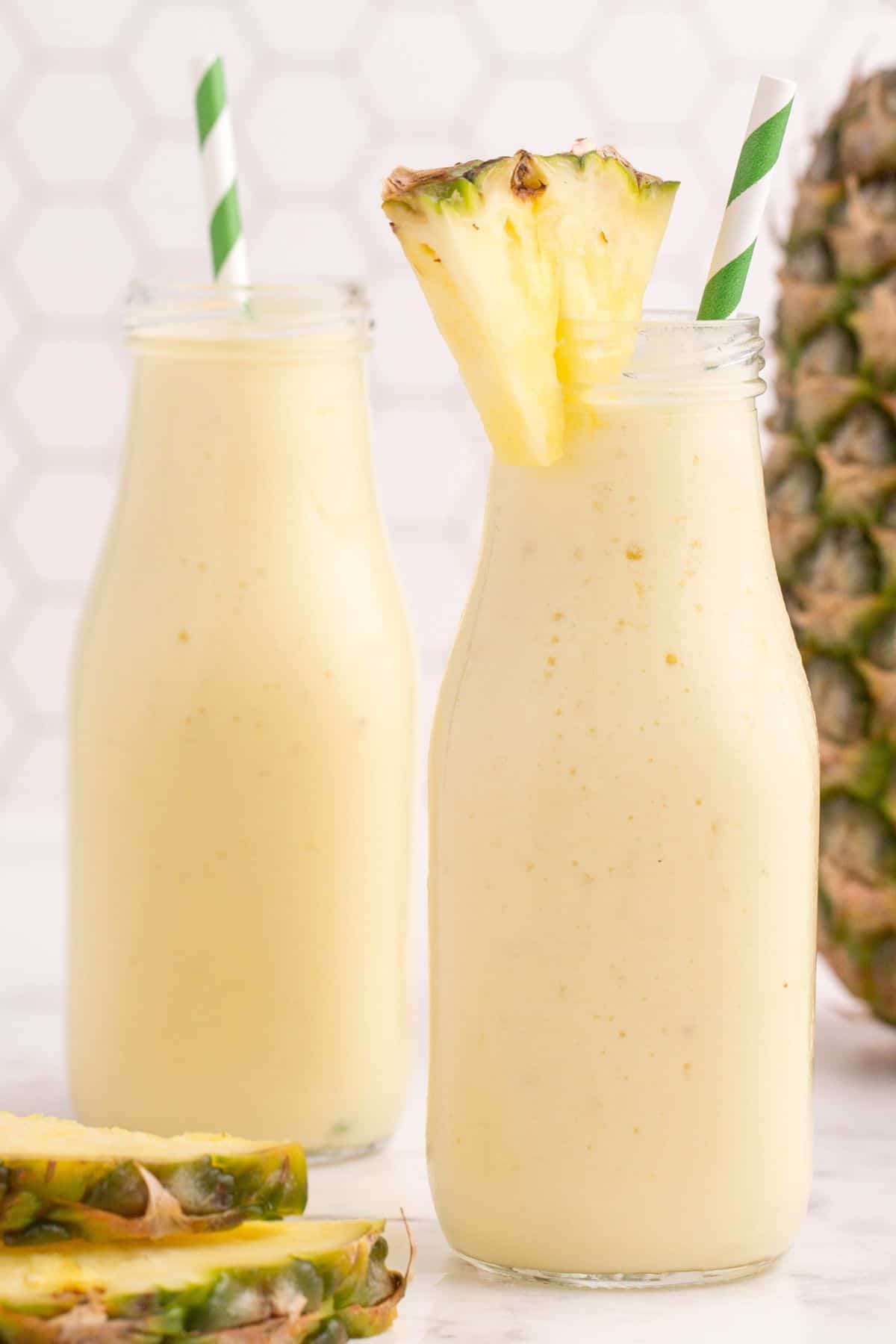 Pineapple smoothies in milk style glasses. Each glass has a white and green striped straw. One glass has a small pineapple wedge on the rim of the glass.