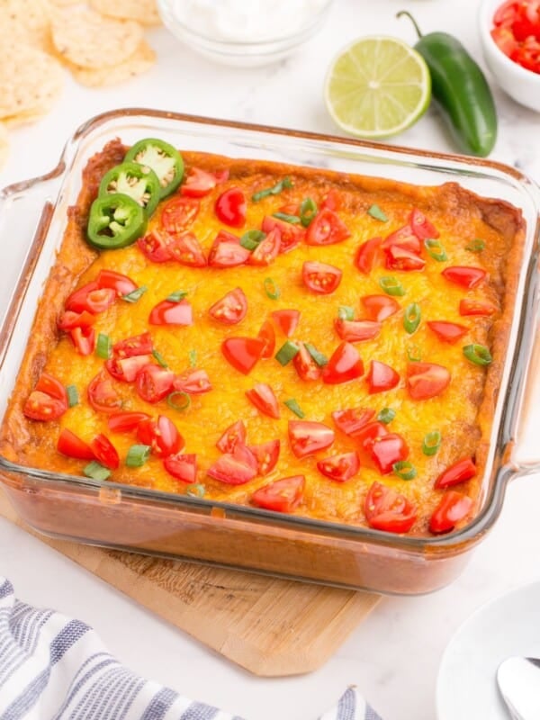 Completed refried bean dip in a glass baking pan. Dip has been garnished with diced tomatoes, green onions, and sliced jalapenos.