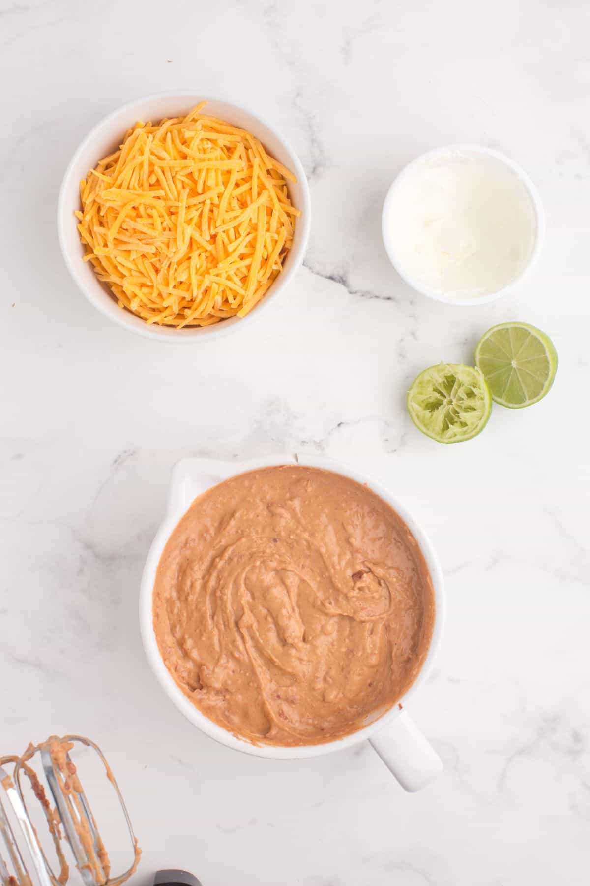 Canned refried beans, sour cream, cream cheese, lime juice, and seasoning mix have been combined with a hand blender in a mixing bowl.