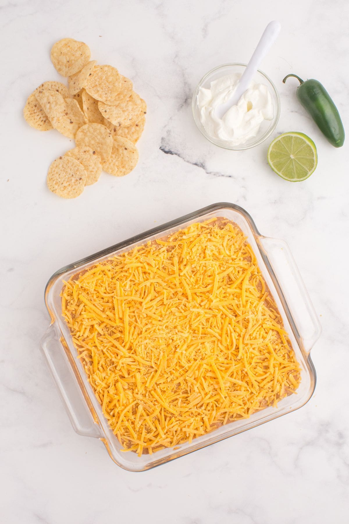 Unbaked refried bean dip is in a glass baking dish and covered with a layer of shredded cheese.
