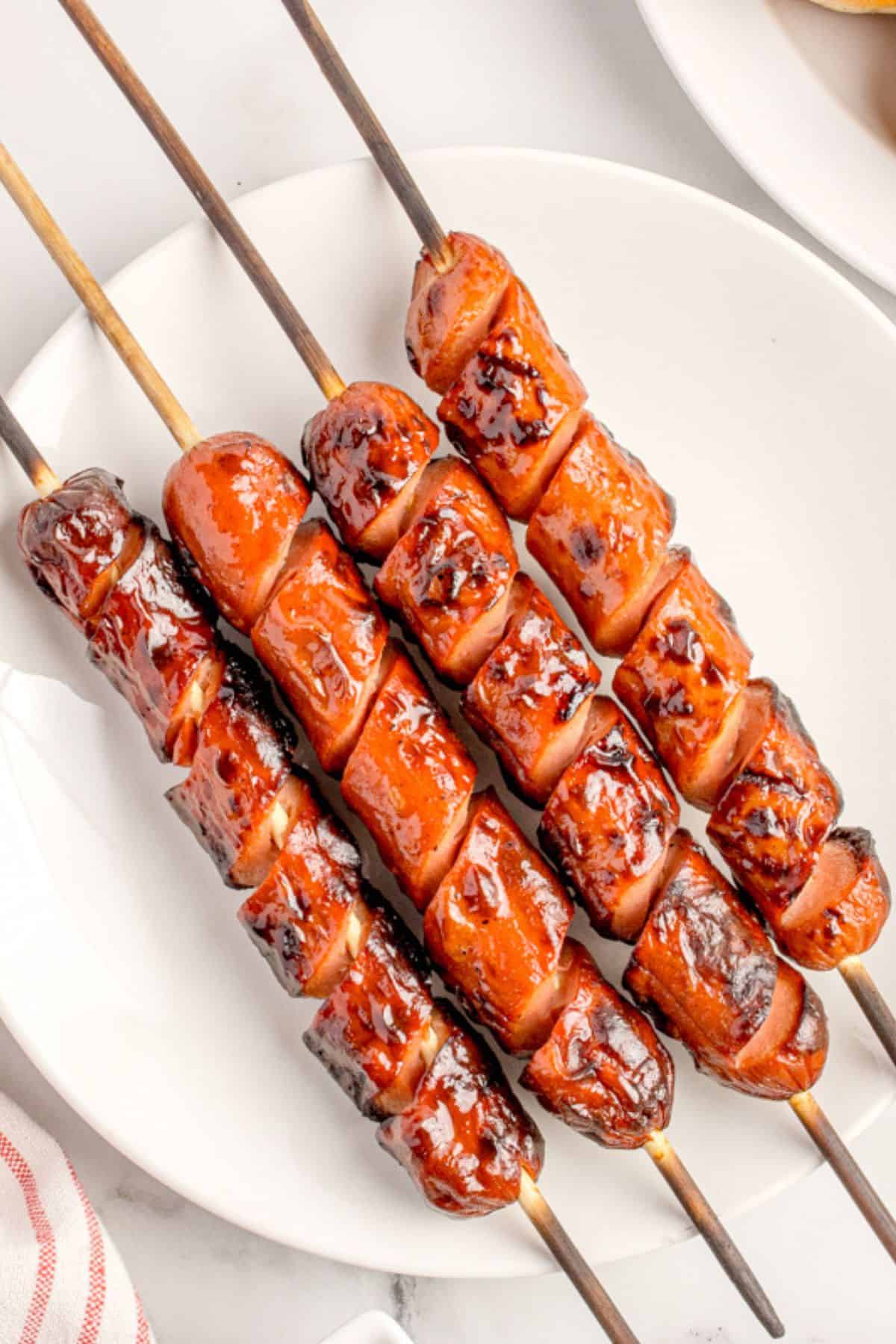 Grilled hot dogs still on the wooden skewers on a plate showing their crispiness on the outside.