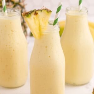 Three milk style bottles filled with pineapple smoothie. Each glass has a white and green striped straw in it and one glass has a small pineapple wedge on the rim.