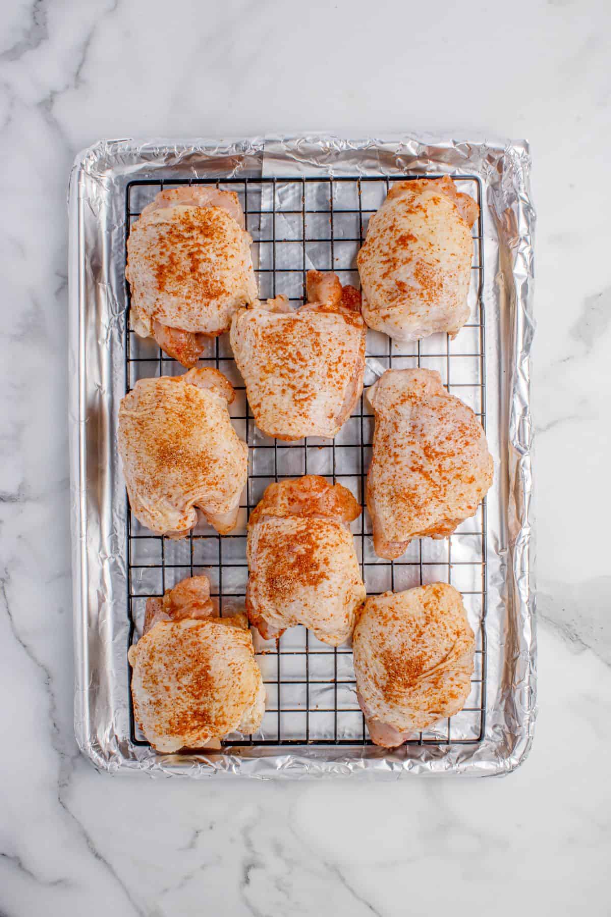 Raw, seasoned chicken thighs on an aluminum foil lined baking sheet with a rack.