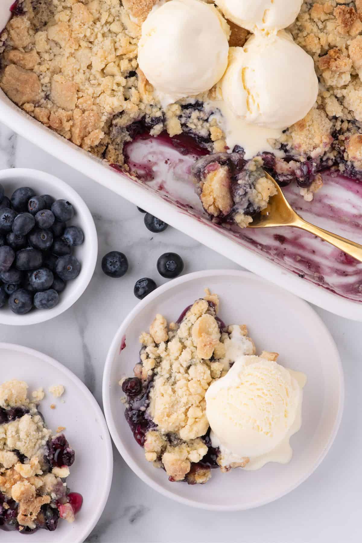 Completed blueberry cobbler in a white baking dish next to two servings of blueberry cobbler in white bowls.