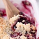 A wooden spoon scooping up a serving of blueberry cobbler out of a white baking dish.