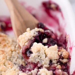 A wooden spoon scooping up a serving of blueberry cobbler out of a white baking dish.