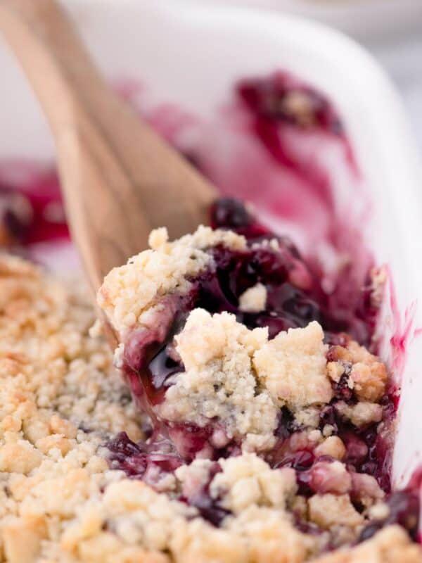A wooden spoon scooping up a serving of blueberry cobbler out of a white baking dish.