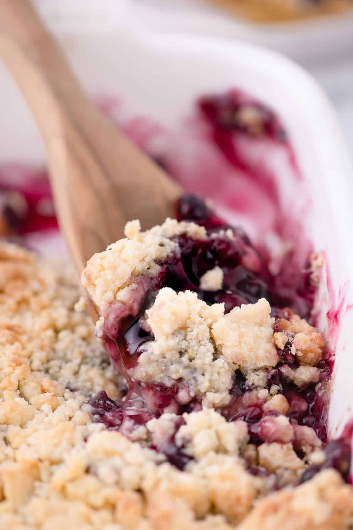 A wooden spoon scooping up a serving of blueberry cobbler out of a white baking dish.