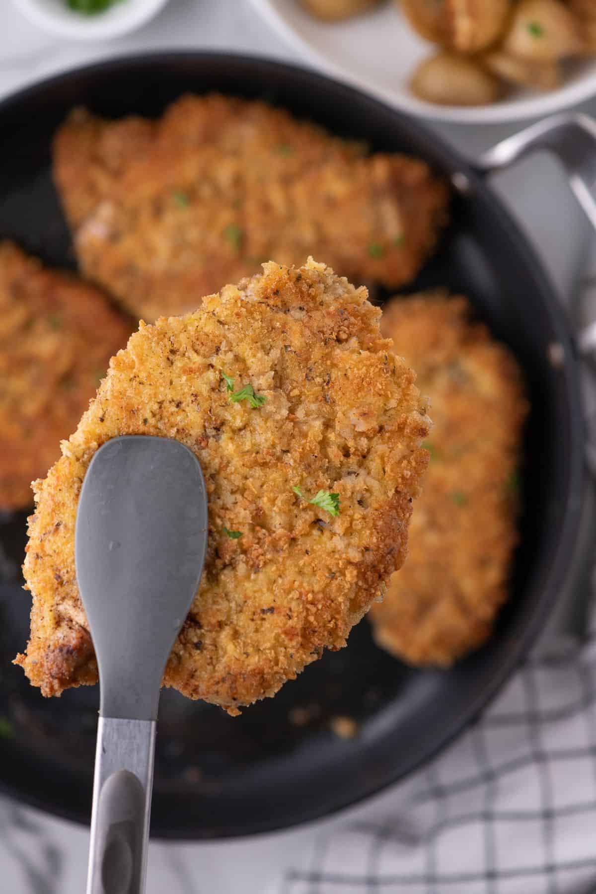 A set of tongs is lifting a pork chop above the skillet with the rest of the pork chops.