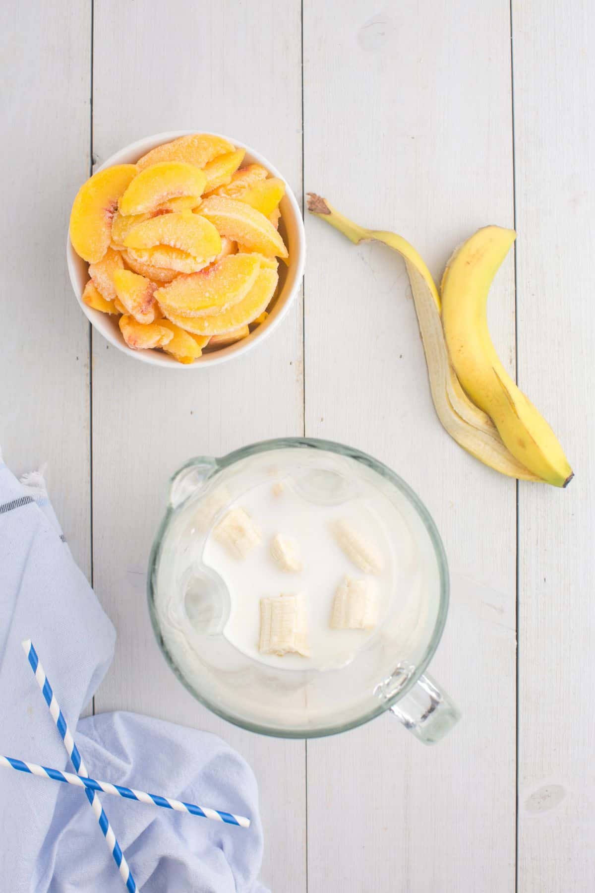 Blender canister with milk, yogurt, and banana in it. A bowl of frozen sliced peaches sits next to the blender canister.