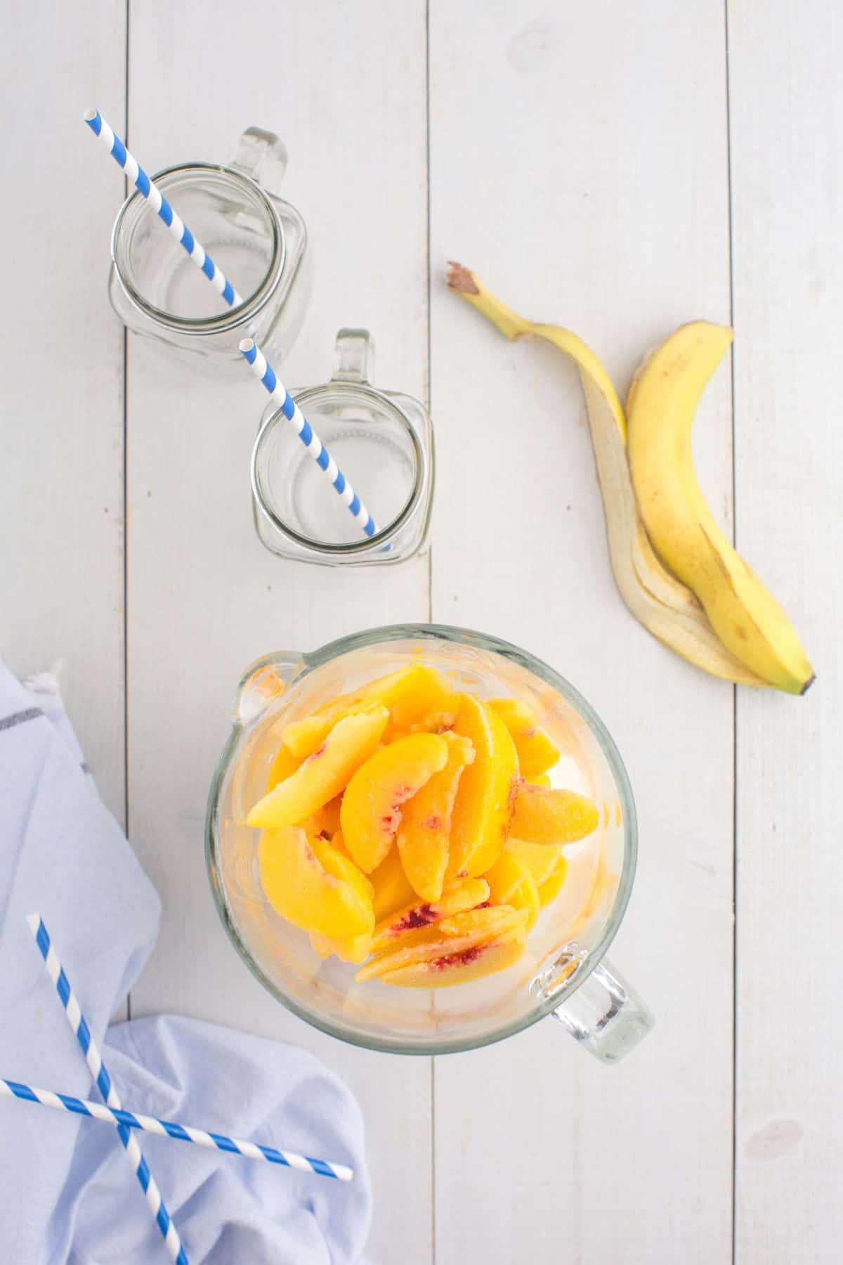 Blender jar layered with milk, yogurt, banana, and sliced peaches. Next to the blender jar is an empty banana peel and two empty glass mugs.