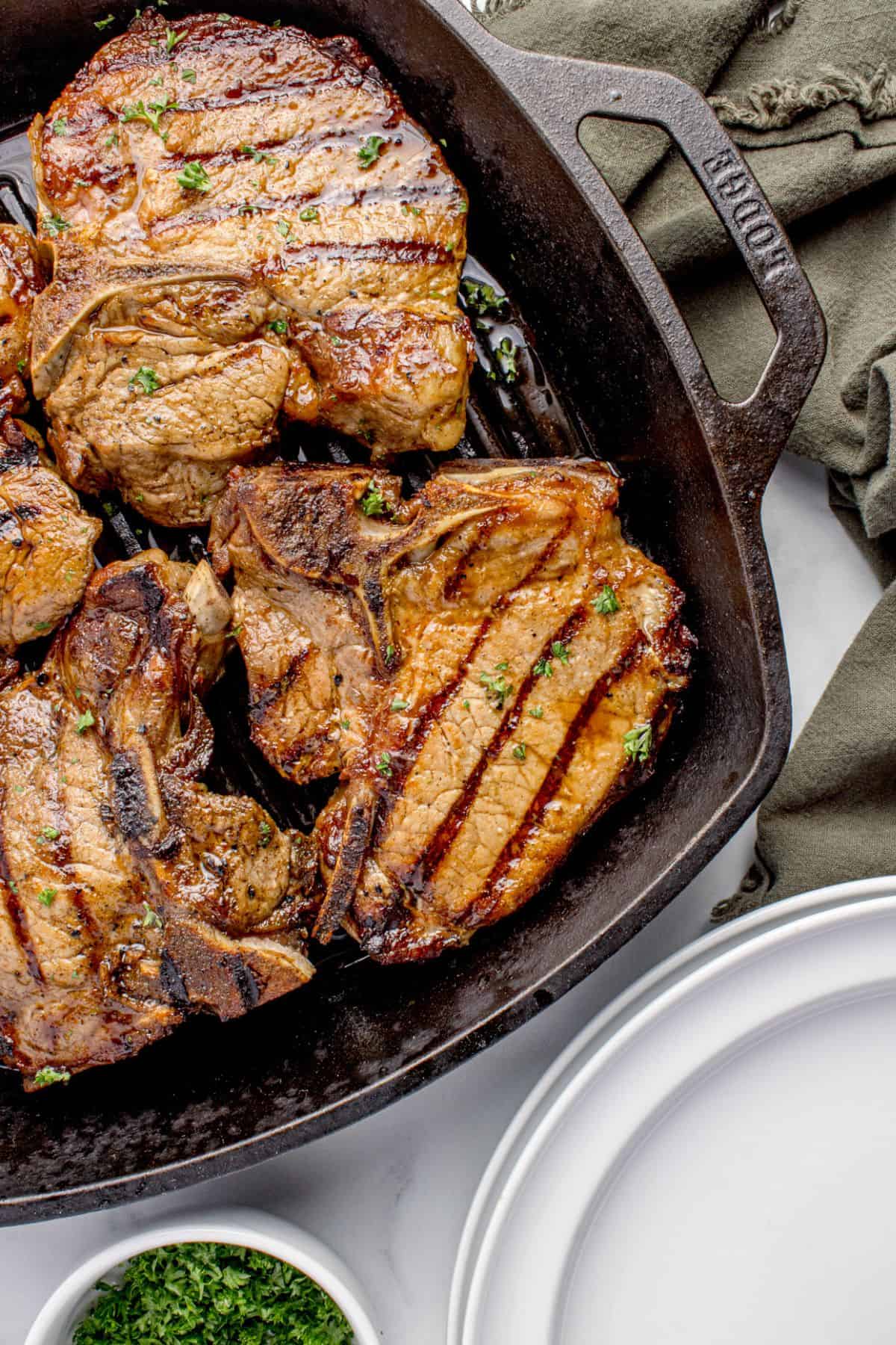 Completed pork chops in a cast iron pan next to a stack of dinner plates and a bowl of parsley.