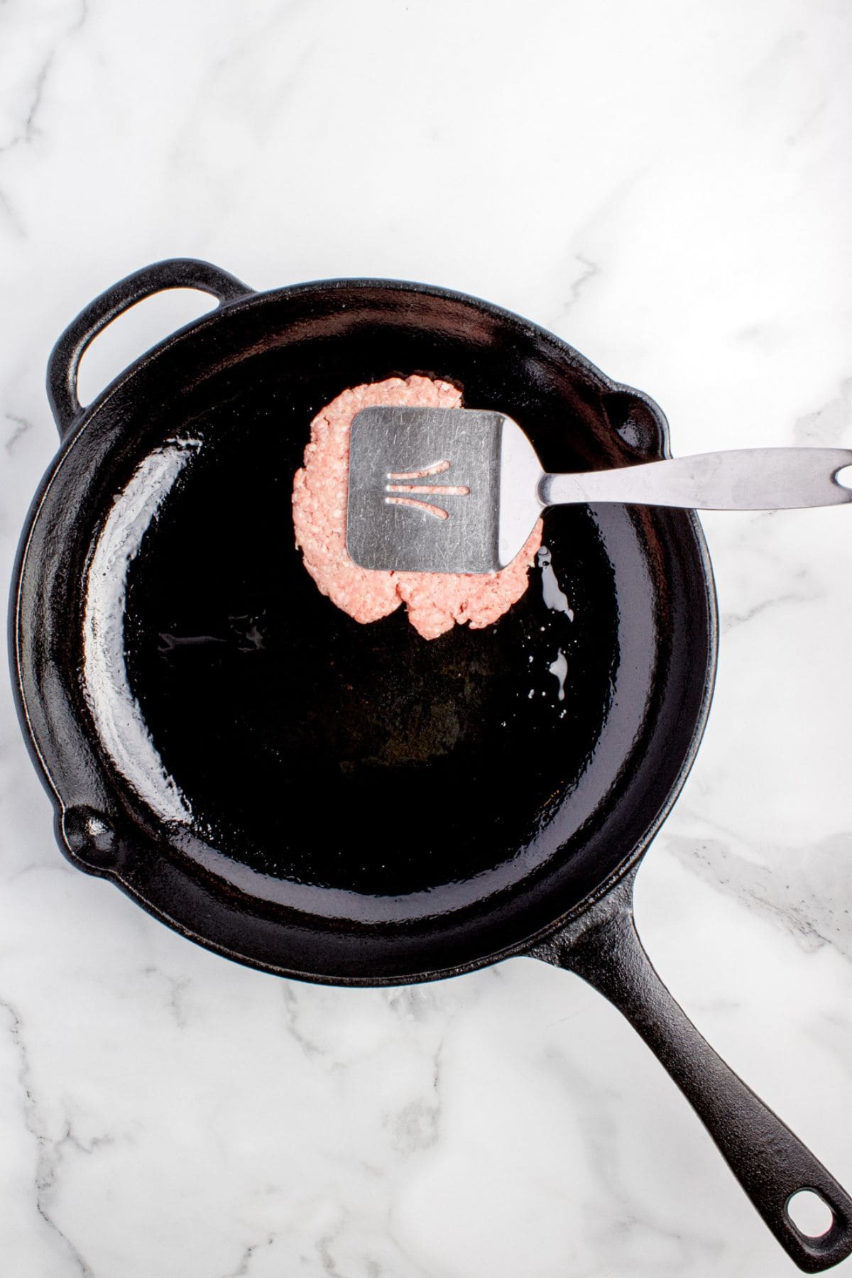 A metal spatula is smashing the burger patty flat in the cast iron pan.